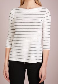 Frau trägt ein weißes Langarmshirt mit schwarzen horizontalen Streifen und schwarze Hosen, steht vor einem schlichten hellen Hintergrund.