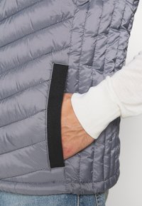 Gepolsterte Daunenweste in Grau mit gestreiftem Muster, ausgestattet mit einer schwarzen Tasche und kontrastierenden weißen Ärmelbündchen. Die Textur wirkt weich und plüschig.