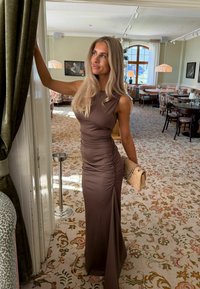 Femme en robe marron sans manches tenant un sac à main beige, debout près d'une porte dans une pièce élégante avec un tapis floral et des meubles classiques.
