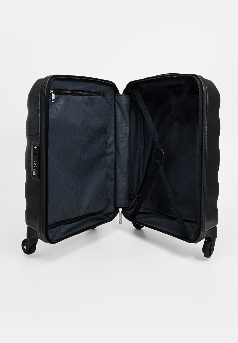 Valise rigide noire avec quatre roues, ouverte pour révéler un intérieur sombre comprenant des poches zippées et un système de sangles croisées pour sécuriser les objets.
