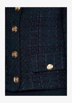 Zwarte textuurjack met een ruitpatroon, voorzien van gouden knopen en een zak. De stof heeft een geweven uitstraling met variërende diktes.