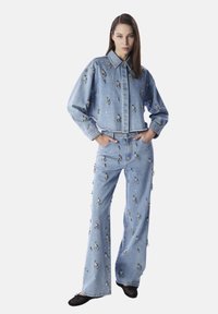 Giacca di jeans azzurro chiaro e pantaloni a zampa d'elefante, entrambi con decorazioni metalliche. La giacca è corta con colletto, e i pantaloni hanno una vita alta.