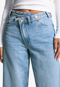 Personne portant un jean en denim bleu clair avec une taille à double bouton unique et un haut court à manches longues blanc.