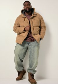 Hombre de pie frente a un fondo simple, vestido con una chaqueta aislante marrón, una camisa burdeos, jeans holgados y desgastados, y botas marrones con cordones.