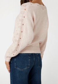 Helle rosa Strickjacke mit Rundhalsausschnitt, Durchbrüchen an den Ärmeln und einem figurbetonten Taillenbund, kombiniert mit dunkelblauen Jeans.