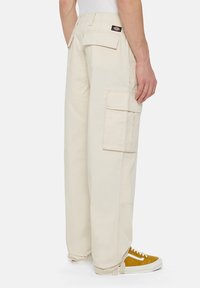 Beige cargobroek met een losse pasvorm, voorzien van zijzakken, een achterzak en een logo patch. Gecombineerd met gele sneakers voor contrast.