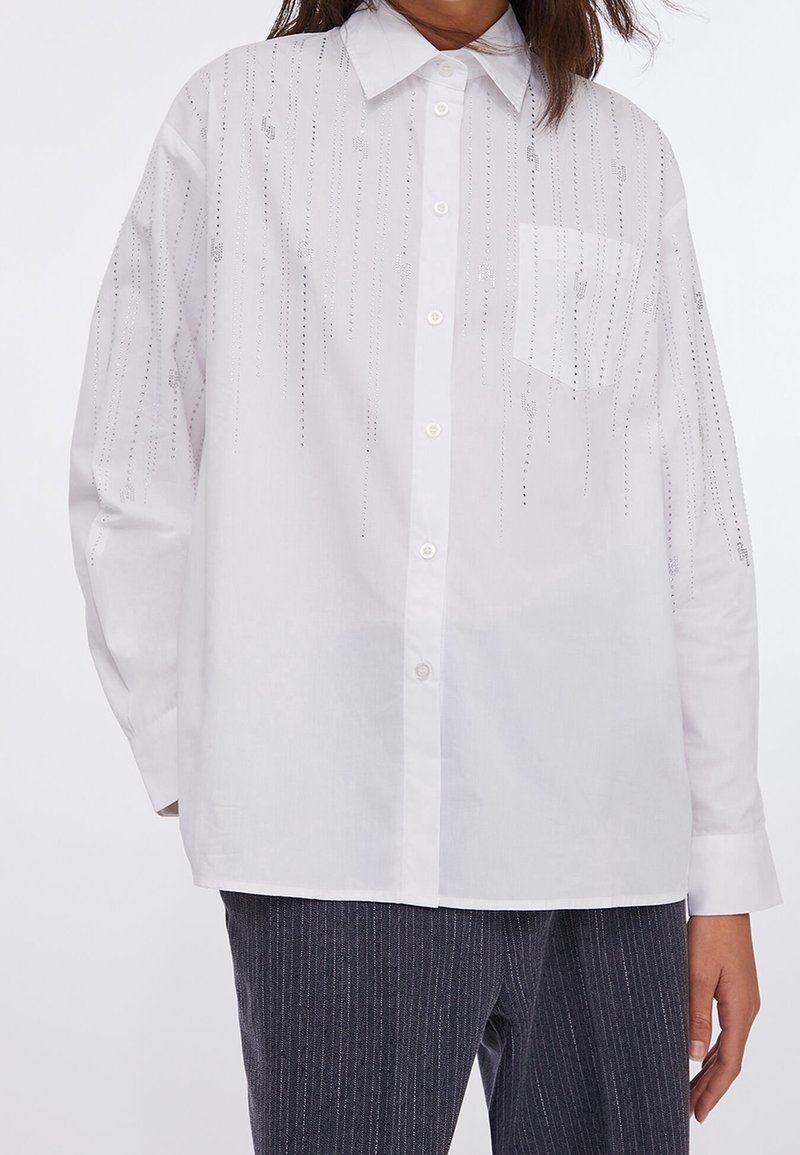 Personne portant une chemise blanche à boutons avec des ornements verticaux en perles argentées et une poche poitrine, assortie à un pantalon sombre à fines rayures.