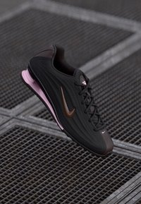 Crne i ružičaste Nike tenisice s smeškim detaljem, vezice vezane, prikazane kako lebde iznad metalne rešetkaste površine.