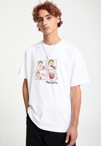 Hvid bomulds-T-shirt med et grafik af fire tegneserieansigtet med lyserøde baggrunde og sort tekst, der siger "VOLCOM." Korte ærmer, løs pasform.