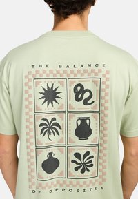 Světle zelené bavlněné tričko s grafickým designem, který obsahuje šest ilustrovaných čtverců v černé a broskvové barvě, s nápisem "OVLIVNĚNÍ PROTIKLADY."