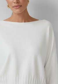 Pull en tricot blanc avec un col large et une texture côtelée sur la moitié inférieure. Doté de manches longues et d'un tissu doux.