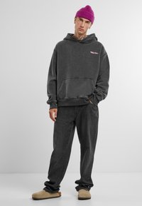 Dunkelgrauer Hoodie mit Fronttasche und pinkem Logo. Getragen mit locker sitzenden dunkelgrauen Jeans und beigen Hausschuhen. Wollige rosa Mütze.