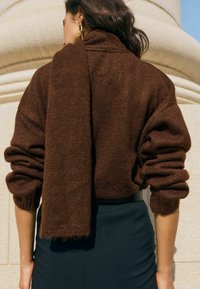 Ruskea neulottu neulepaita käännetyin hihoin ja siihen sopiva kaulaliina olkapäällä, yhdistetty mustaan vartalonmyötäiseen hameeseen.