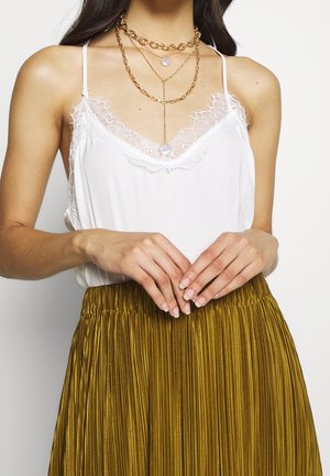 Camisole blanche avec dentelle, superposée sous des colliers multi-chaînes dorés ; associée à une jupe plissée en or métallique.