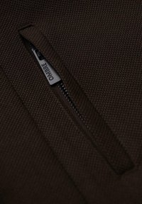 Tejido marrón texturizado con un bolsillo con cremallera que presenta una pestaña negra etiquetada como "OMBRE". Vista en primer plano que resalta el material y el diseño.