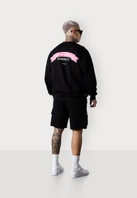Sweatshirt noir avec un texte graphique rose dans le dos, associé à un short cargo noir. Baskets blanches et détail de tatouage visible sur la jambe.