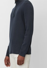 Man som bär en marinblå långärmad stickad tröja med halzzipper, kombinerad med off-white byxor mot en enfärgad bakgrund.