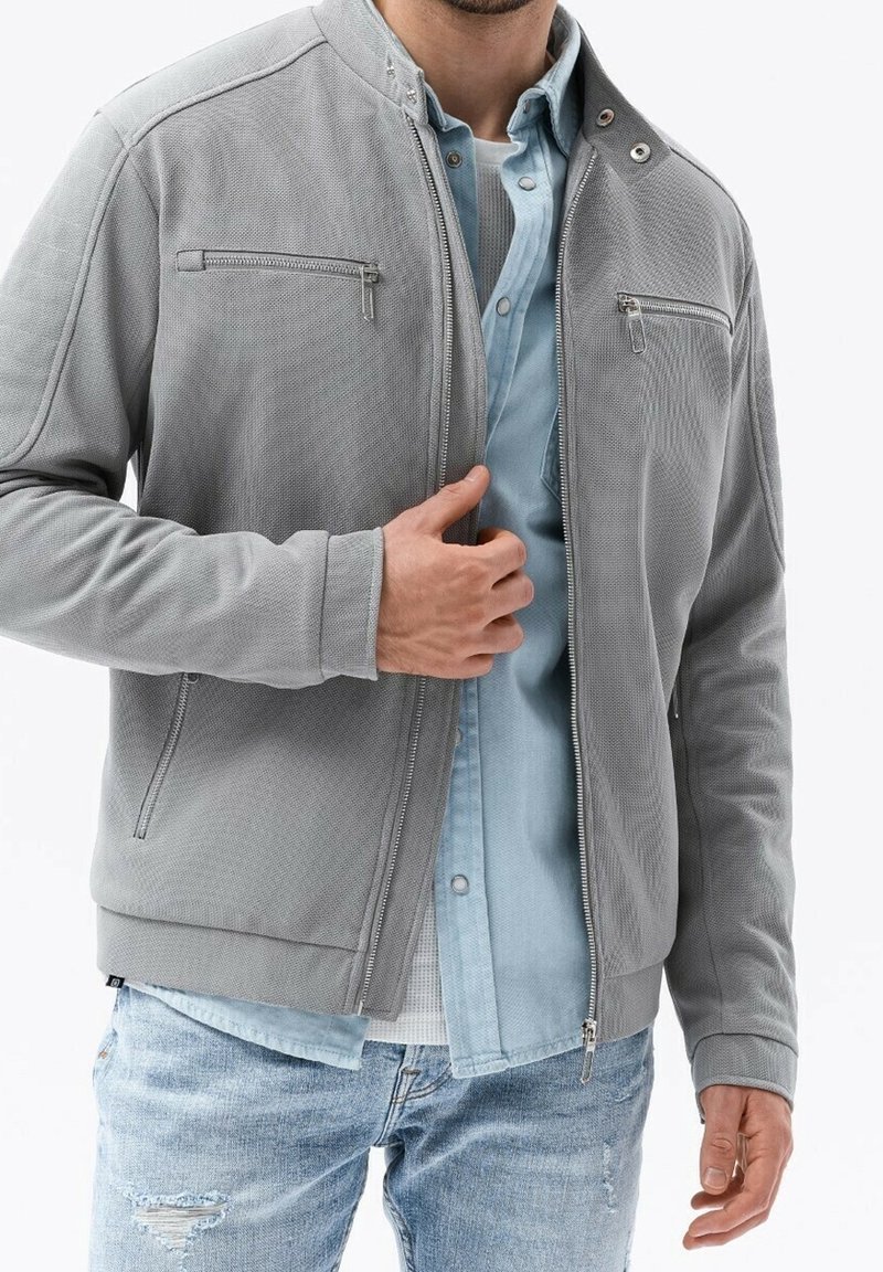 Grijze textuurjacket met een ritssluiting, twee ritsborstzakken en een staande kraag. Draagt over een lichtblauw shirt.