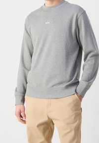 Hombre que lleva un sudadera de manga larga gris claro con el texto "BOSS" en el pecho y pantalones beige, con una mano en el bolsillo, contra un fondo blanco.