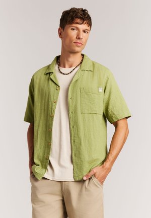 Hellgrünes, strukturiertes Kurzarm-Hemd mit Knöpfen und Brusttasche, mit braunen Knöpfen, getragen über einem neutralfarbenen T-Shirt und beigen Hosen.
