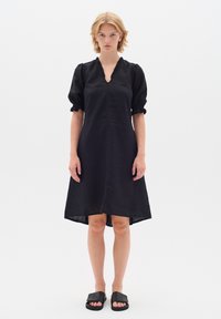 Schwarzes Leinenkleid mit V-Ausschnitt, gerafften kurzen Ärmeln und einem knielangen Saum. Glatte Textur mit dezenten Designdetails.