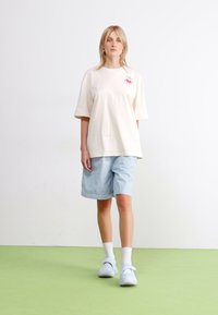 T-shirt oversize in cotone crema con maniche corte e design floreale rosa. Abbinato a pantaloni corti a righe blu e calzature azzurro chiaro.