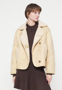 Veste en shearling dans un beige clair, avec un large col, un devant double boutonnage et deux poches latérales. Texture extérieure lisse, doublure intérieure douce.
