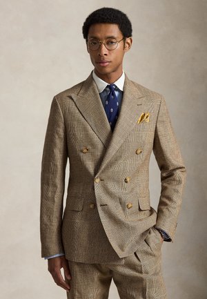 Bărbat purtând un costum dublu bej în carouri cu cravată bleumarin cu model, batistă galbenă în buzunar, ochelari rotunzi și o mână în buzunar.