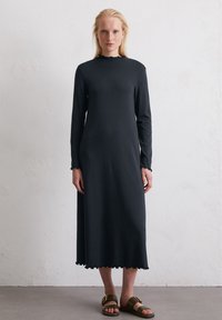 Czarna sukienka z długim rękawem i okrągłym dekoltem, z falbaniastym wykończeniem na rękawach i u dołu, wykonana z gładkiego materiału, zestawiona z prostymi brązowymi sandałami.