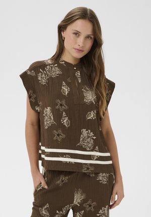 Kobieta ubrana w brązowy bezrękawnik i spodnie z białymi wzorami morskim tematycznymi oraz dwoma poziomymi białymi paskami na bluzce.