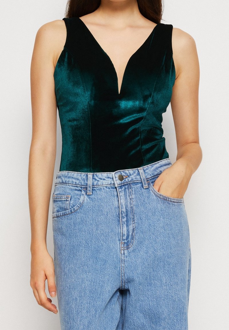 Woman wearing sleeveless dark green velvet top with deep V neckline tucked into high-waisted light blue denim jeans, hand in pocket.