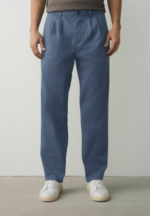 Mann trägt blaue plissierte Hose und weiße Turnschuhe, steht vor einer schlichten hellgrauen Wand und einem Betonboden.