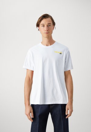 Camiseta de algodón blanca de manga corta, con cuello redondo, que presenta el texto amarillo "Lacoste" y un logo de cocodrilo verde en la parte izquierda del pecho.