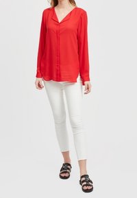 Blusa rossa a maniche lunghe con scollo a V e chiusura con bottoni, abbinata a pantaloni bianchi aderenti e sandali neri con cinturini.