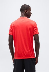 Άνδρας με πλάτη φορώντας φωτεινό κόκκινο αθλητικό μπλουζάκι κοντομάνικο και μαύρο σορτς σε λευκό φόντο.