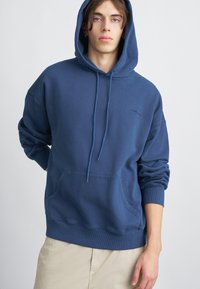 Bluza z kapturem w kolorze granatowym. Materiał bawełniany, luźny krój, kieszeń kangurza, ściągacze przy mankietach i u dołu, z delikatnym logo na klatce piersiowej.