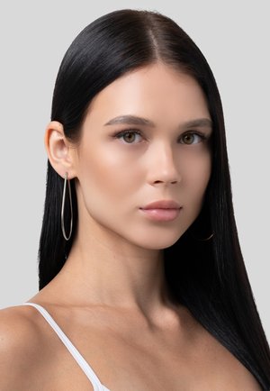 Giovane donna con lunghi capelli neri, indossa un top bianco a spalline sottili e grandi orecchini a cerchio d'argento, guarda direttamente davanti a uno sfondo grigio.