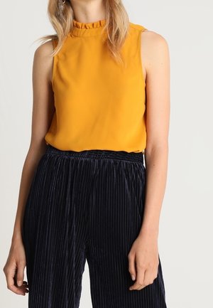 Femme portant un chemisier sans manches jaune moutarde avec un col à volants et un pantalon noir plissé en velours taille haute.