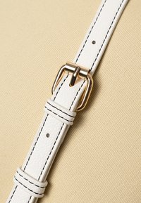 Weißes strukturiertes Lederarmband mit schwarzem Nähen, ausgestattet mit einer goldenen Metallschnalle. Der Hintergrund ist aus beigem Stoff.