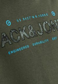 Camiseta de algodón color verde oliva con texto en relieve en negro que dice "JACK & JONES" con acentos azules y coordenadas geográficas.