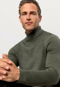 Grüne Rollkragenpullover mit weicher Textur und gerippten Bündchen. Nahaufnahme eines Mannes, der die Hände verschränkt, und dabei das taillierte Design des Pullovers hervorhebt.
