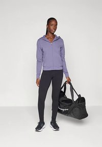 Femme portant un sweat à capuche zippé violet, des leggings noirs et des baskets, tenant un sac de sport noir, se tenant devant un fond clair.
