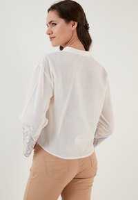 Blouse en lin blanc à manches longues, ornée d'accents brodés bleu et or sur les poignets. Ourlet arrondi et texture légère.