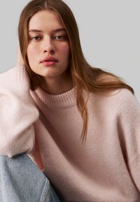 Maglione rosa chiaro, con una morbida trama, dotato di un colletto alto a coste. La modella è seduta in modo informale, indossando jeans blu e ha i capelli lunghi e lisci.