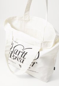 Vit canvas tote bag med långa handtag, med svart text och logotypdesign på en sida, samt ett rymligt, ofodrat inre.