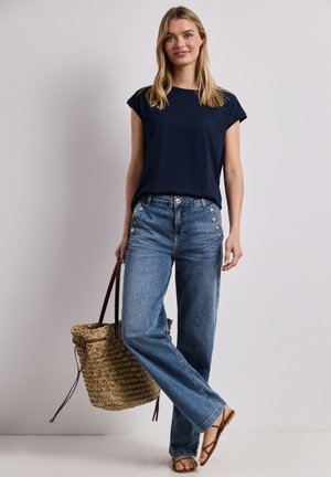 Kobieta w granatowej bluzce, niebieskich jeansach z wysokim stanem i guzikami, brązowe sandały, nosząca tkany słomkowy worek, stojąca na jednolitym tle.