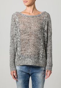 Maglione leggero e a maglia aperta in grigio e nero con scollatura ampia, maniche lunghe e polsini a coste. Indossato con jeans aderenti.