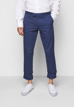 Man met marineblauwe opgerolde chino's, een wit overhemd en witte sneakers, staand tegen een effen lichte achtergrond.