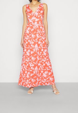 Kvinde iført en koralfarvet maxikjole uden ærmer med blomsterprint i hvid og hvide sandaler med stropper, stående mod en ensfarvet baggrund.