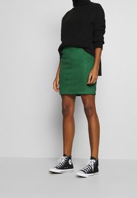 Grön mocka minikjol i kombination med en svart ribbad polotröja och svarta högkänkta sneakers. Outfiten har en avslappnad, lagerpåkostad look.